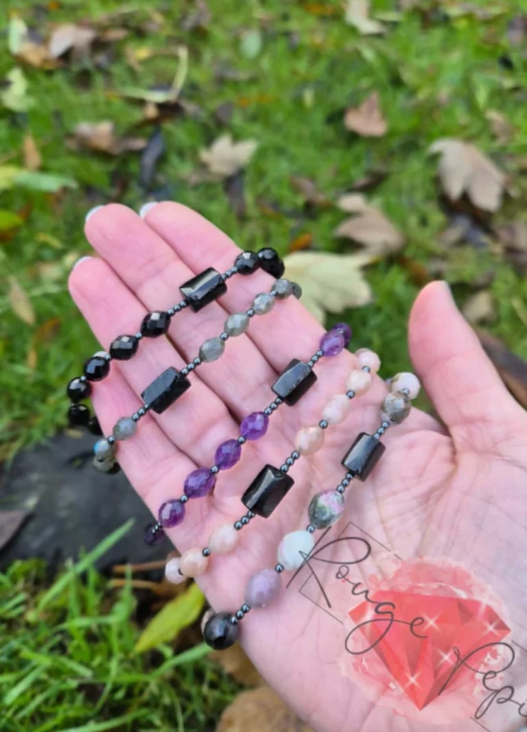 Hand holding five beaded bracelets with black hematite, purple amethyst, and gray stones on grass background